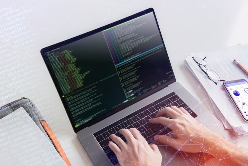 hands typing on the keyboard, coding signs on the laptop screen, phone and glasses on the table